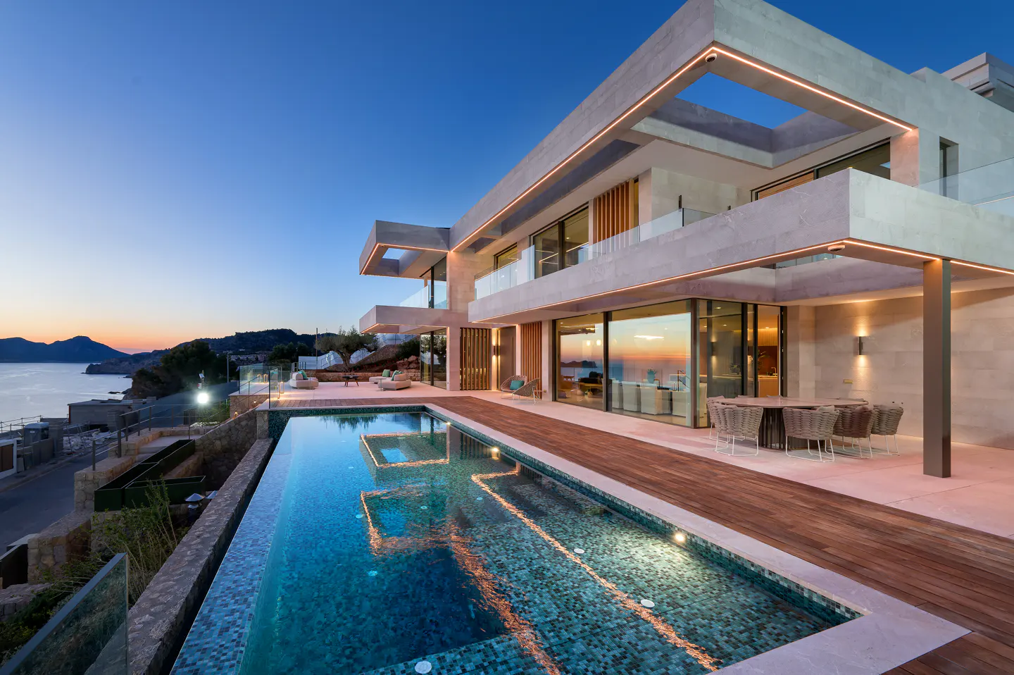 Modern luxury villa with a pool overlooking the ocean at dusk. The villa has a wooden deck and outdoor dining area.