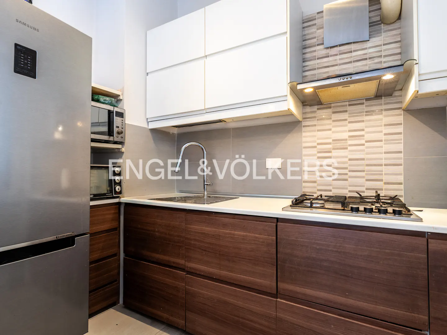A modern kitchen with stainless steel fridge, wood cabinets, white countertops, and a gas stovetop.