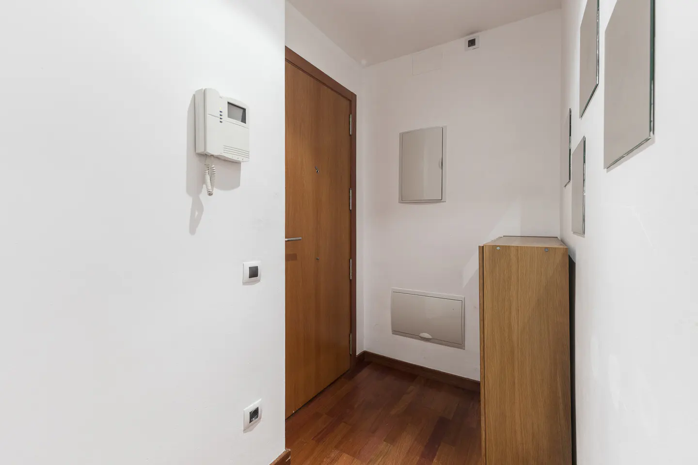 Hallway with white walls, a wood door, and wood floors. An intercom is on the wall, along with a cabinet and mirrors.