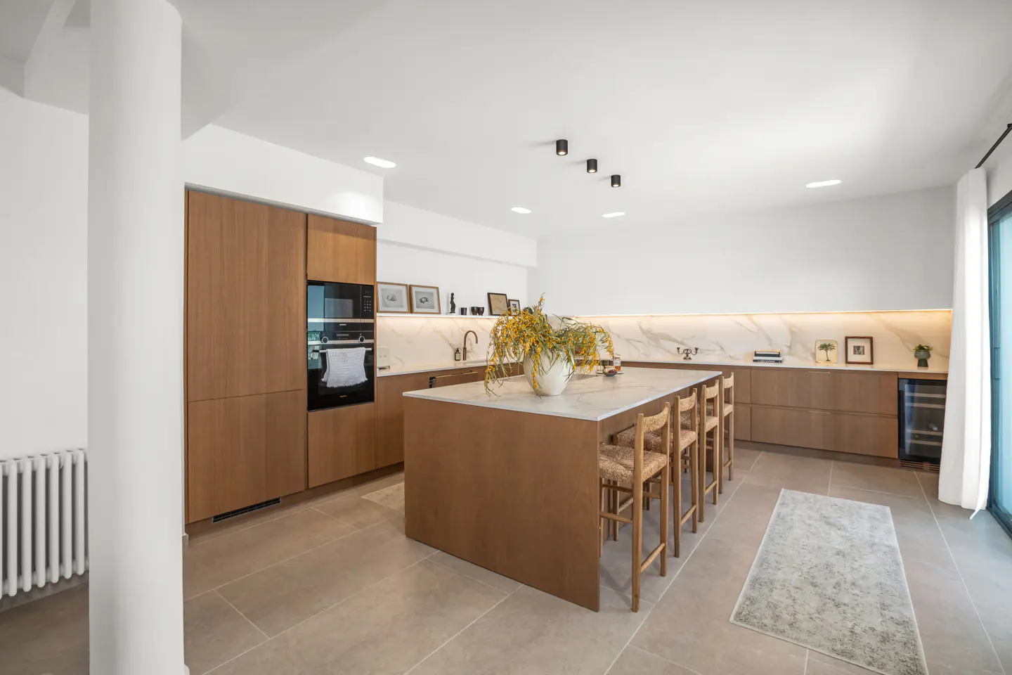Bright, modern kitchen with wood cabinets, marble countertops, and an island with seating. A vase of yellow flowers sits on the island.