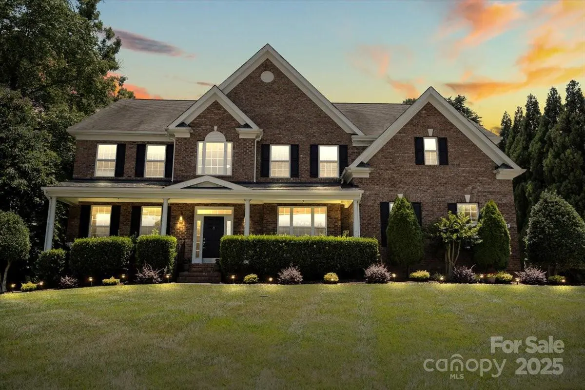 Two-story brick house with black shutters and glowing interior lights at dusk, set behind a manicured lawn and hedges.