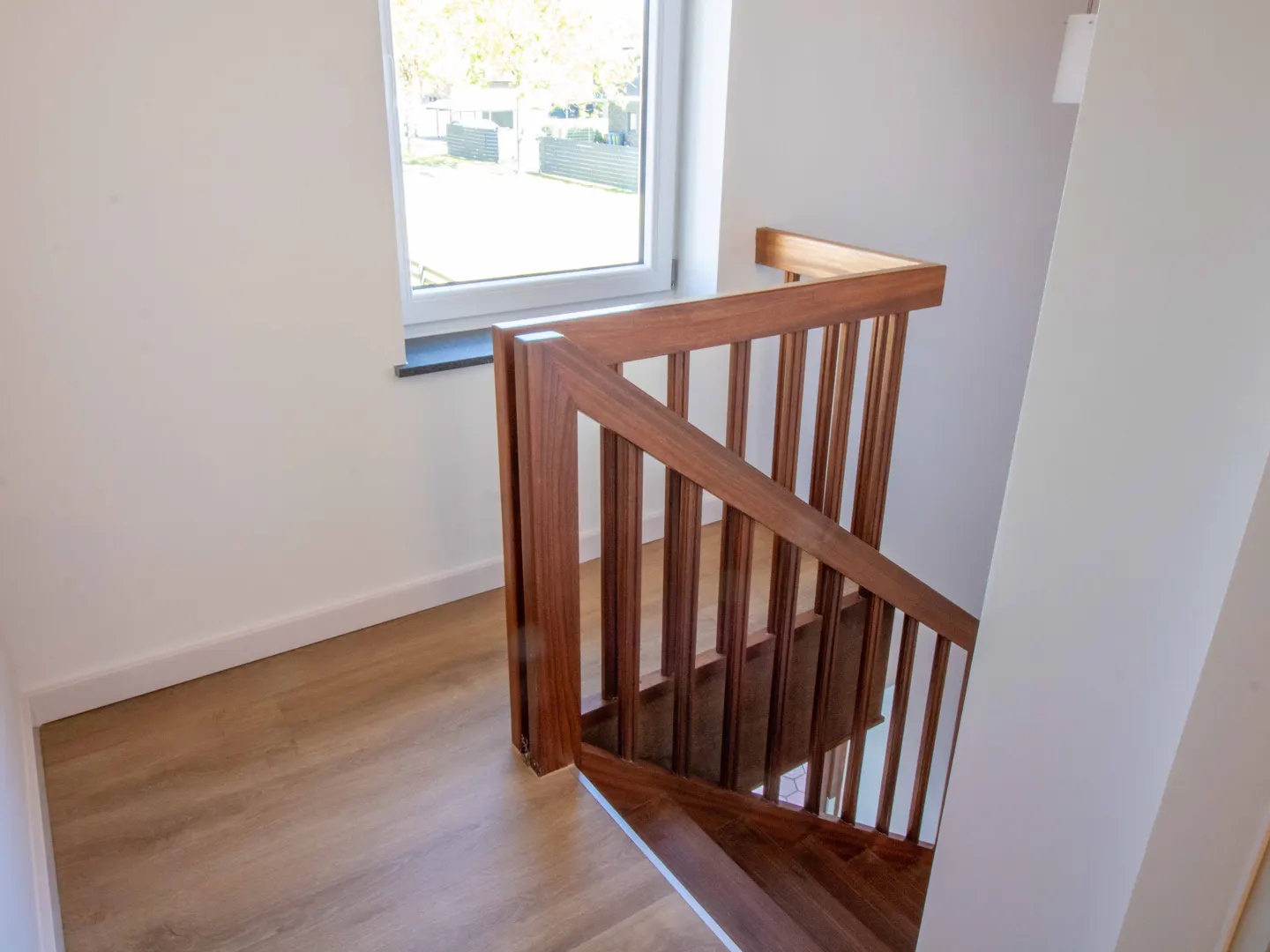 Modern wooden staircase with a sleek railing next to a bright window in a white-walled hallway with light wood flooring.