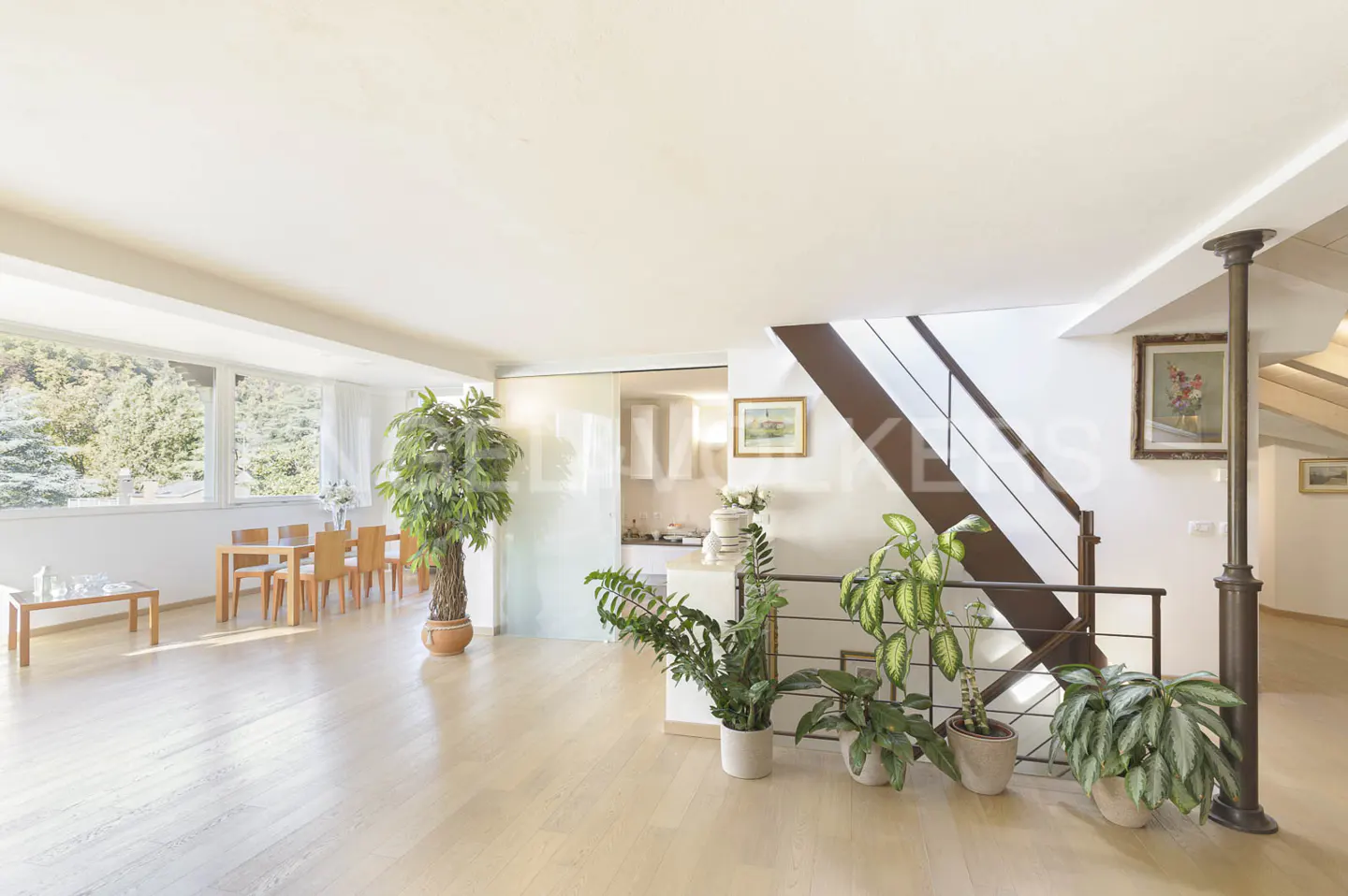 Bright, open-plan living space with light wood floors, dining table, staircase, and potted plants. Large windows offer a view of trees.