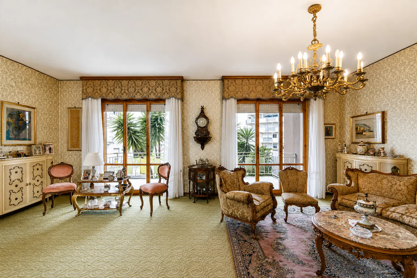 A traditionally decorated living room with patterned wallpaper, ornate furniture, and a large chandelier. Two windows overlook palm trees.