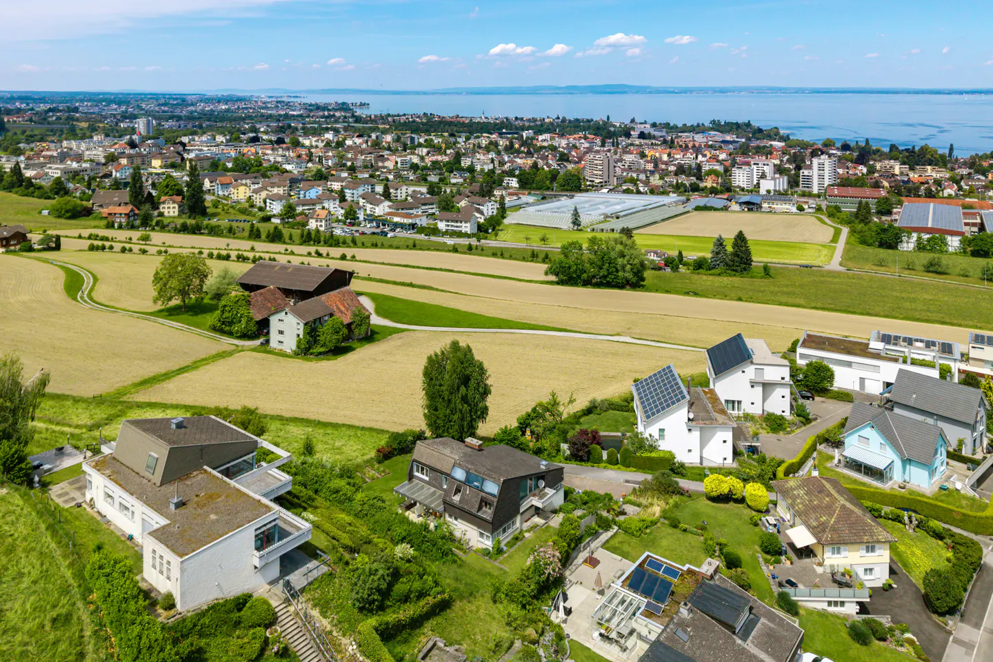 Aerial view of a residential area with modern houses, fields, and a town near a lake under a blue sky.