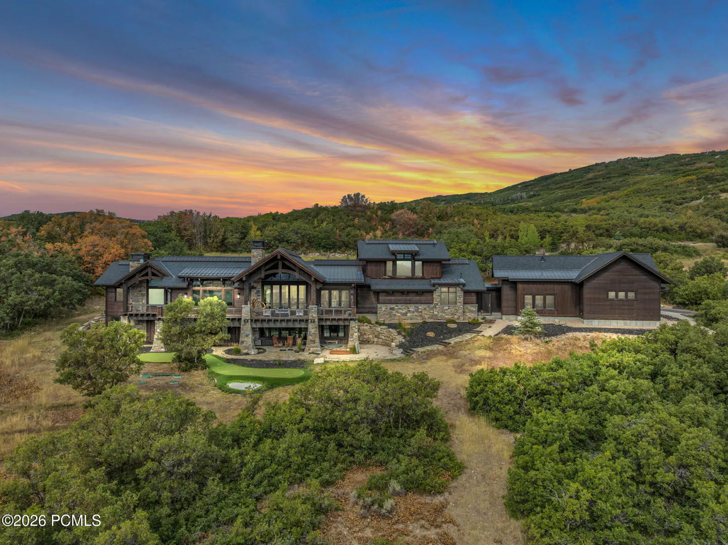 A large, modern brown house with stone accents sits on a hill surrounded by trees, under a colorful sunset sky.