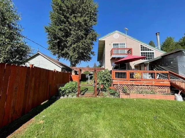 Backyard of a light-colored house with a wooden deck, red umbrella, and a tall wooden fence under a clear blue sky.