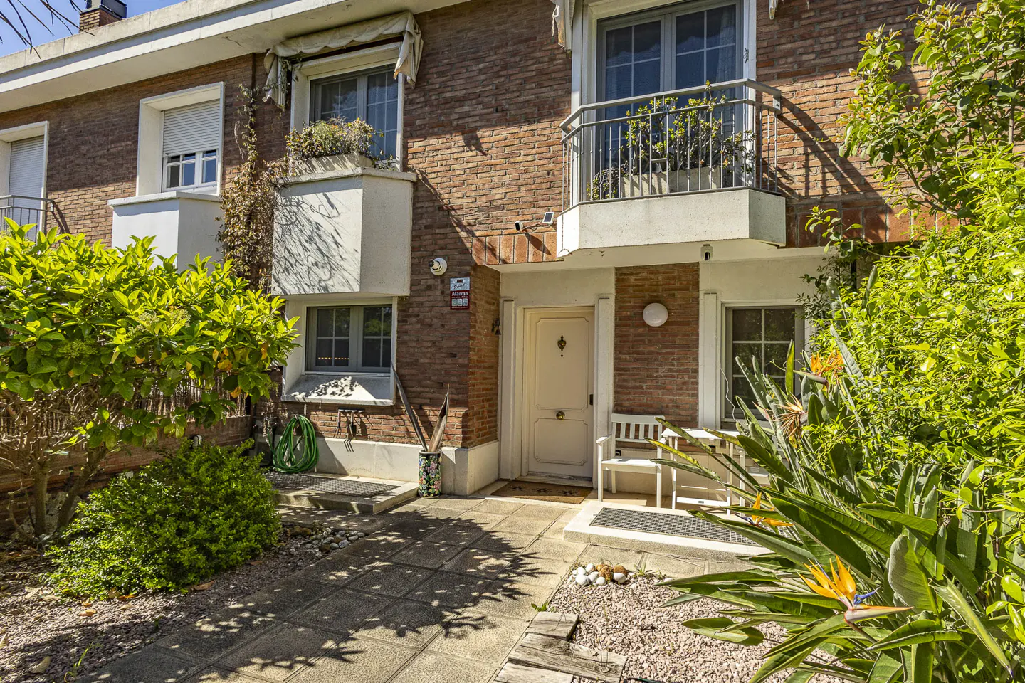 Brick townhouse with a white door, small balcony, and lush greenery in the front yard. A white bench sits near the entrance.