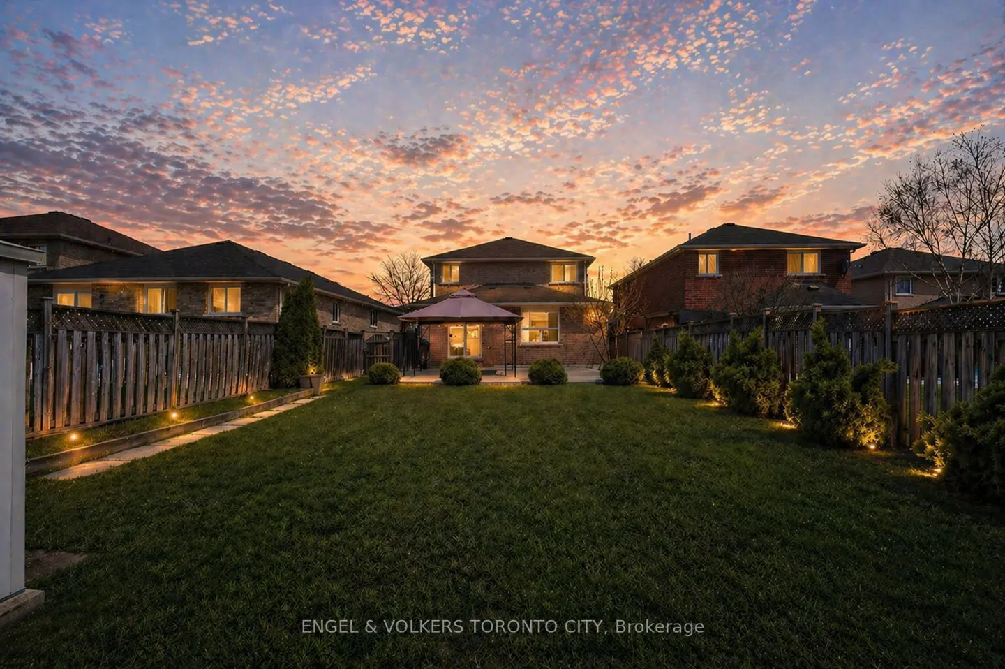 Backyard view of a two-story house with a green lawn, wooden fence, and a gazebo at sunset.