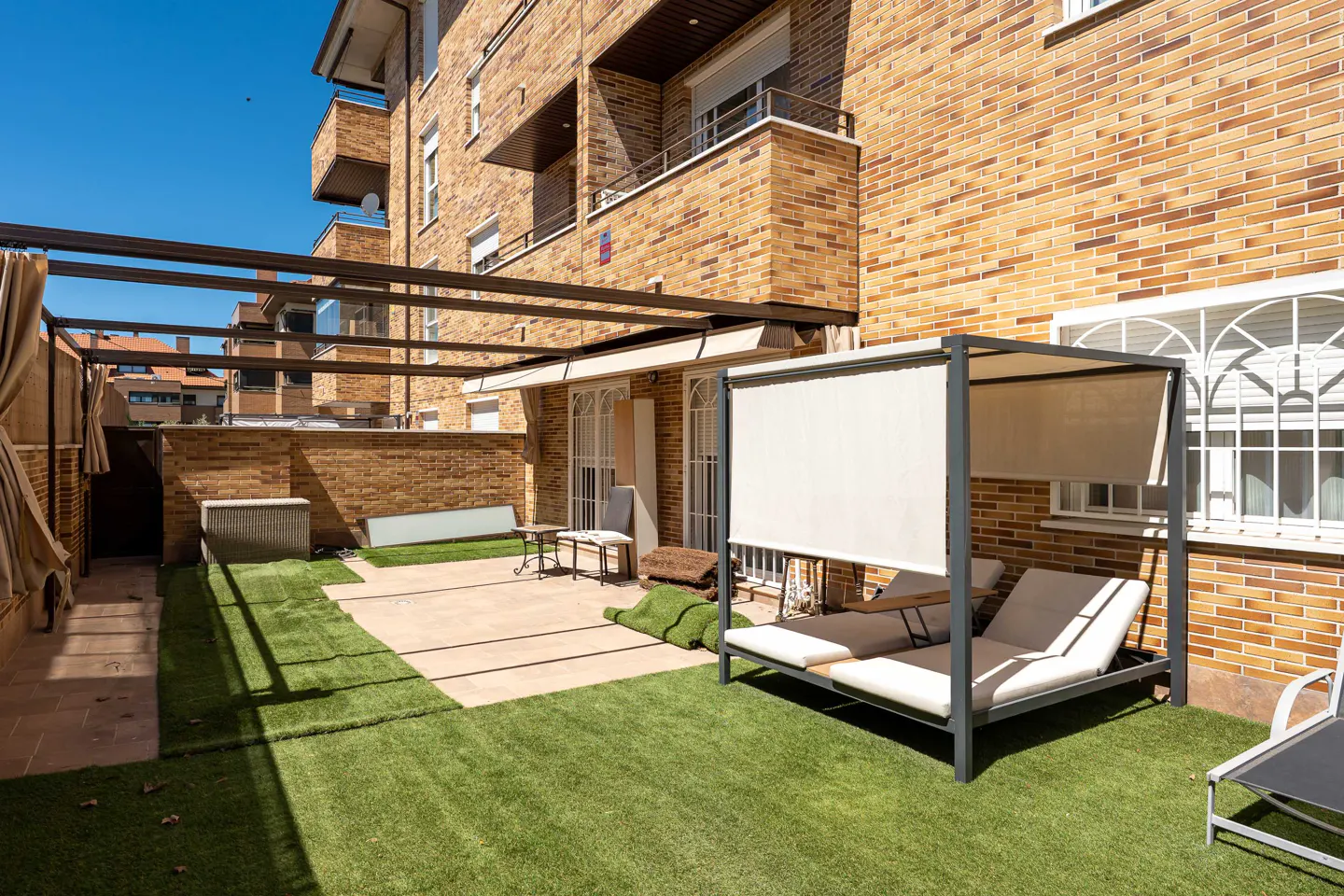Outdoor patio with artificial grass, lounge chairs under a canopy, and a brick building in the background.