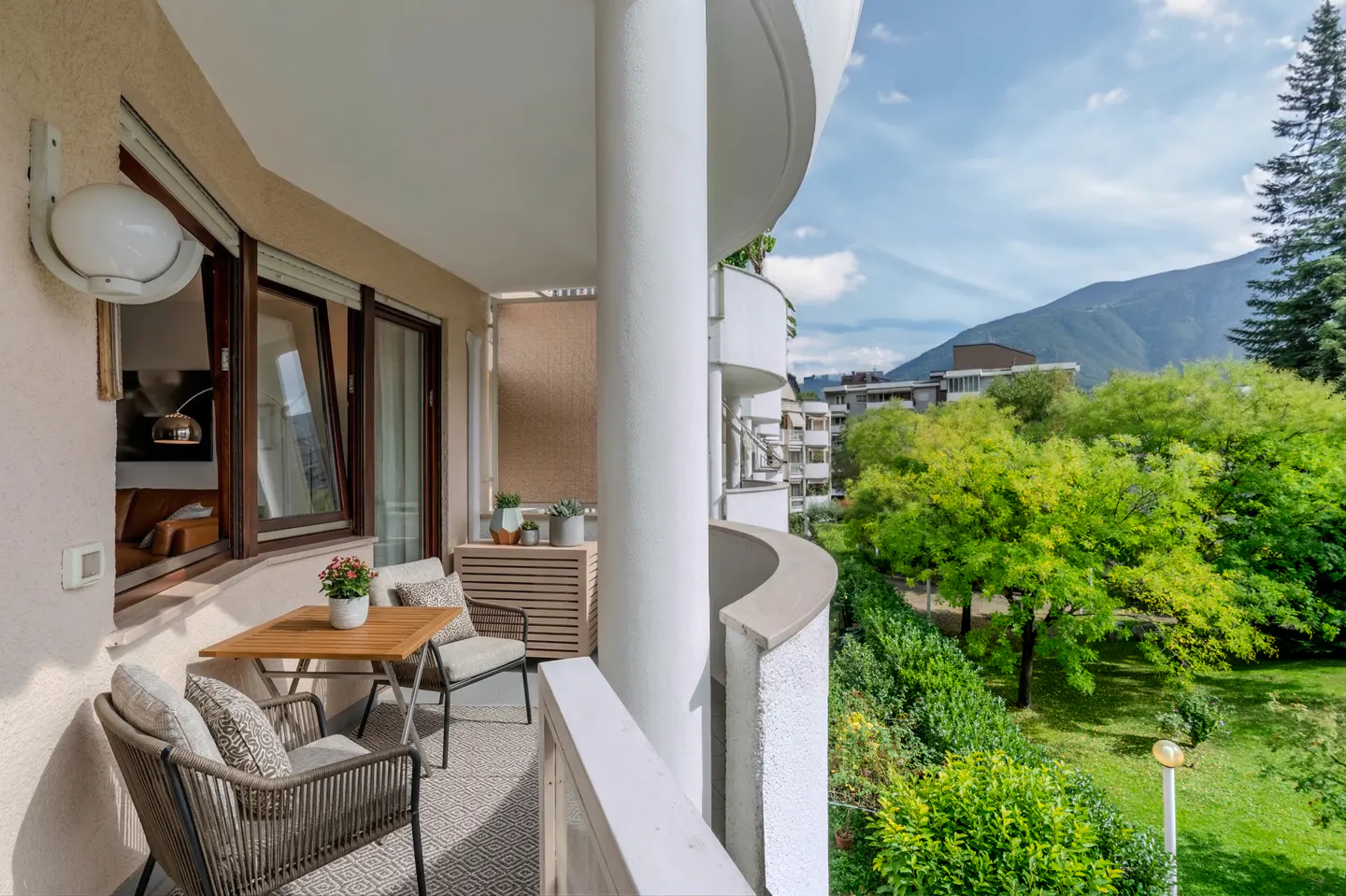 A curved balcony with a table, chairs, and a view of green trees and a mountain.