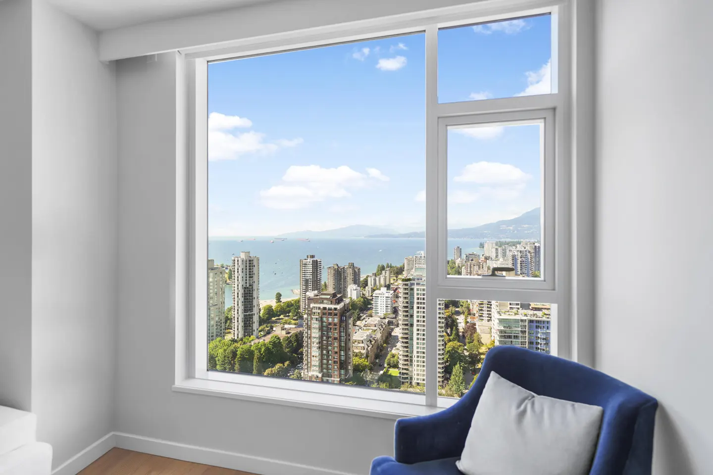 Bright room with a large window overlooking a city skyline, ocean, and mountains. A blue chair with a white pillow sits in the corner.