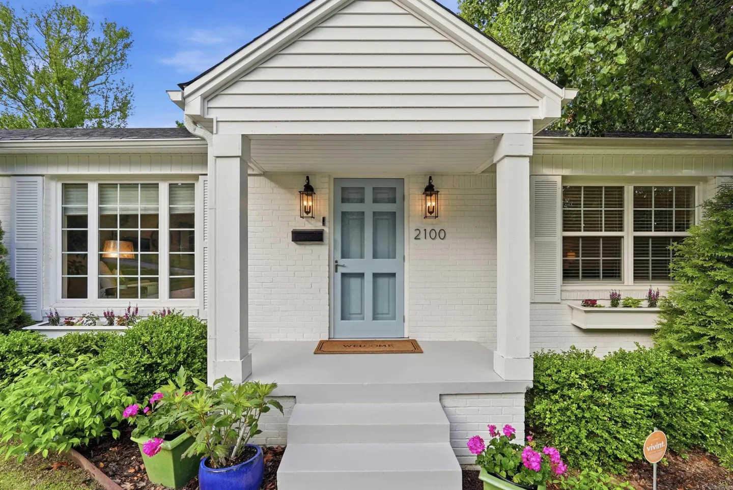 Front view of a white brick house with a light blue door and white pillars. The house number "2100" is visible above the door. Green bushes and colorful potted plants surround the entrance.