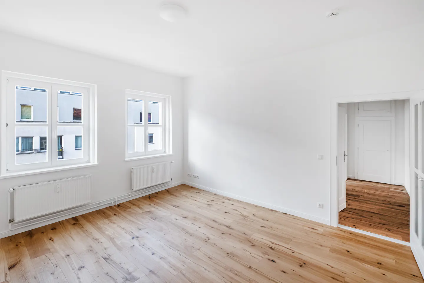 Bright, empty room with hardwood floors, white walls, and two windows. A doorway leads to another room with similar features.