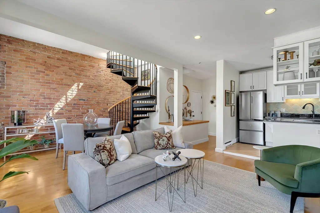 Open-concept living space with exposed brick wall, spiral staircase, grey sofa, and white kitchen with stainless steel appliances.