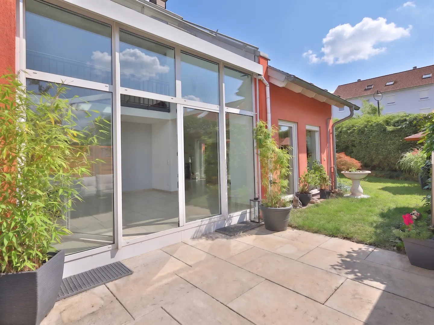 Exterior view of a modern home with a large glass sunroom, a stone patio, and a green lawn.