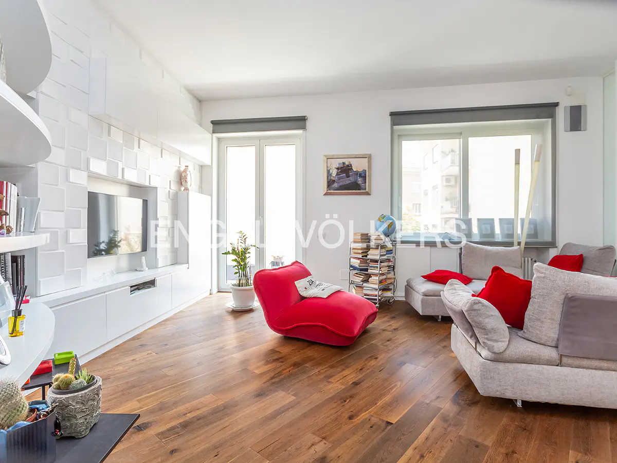Living room with hardwood floors, white walls, and a gray sofa with red pillows. A red chaise lounge sits near a window.