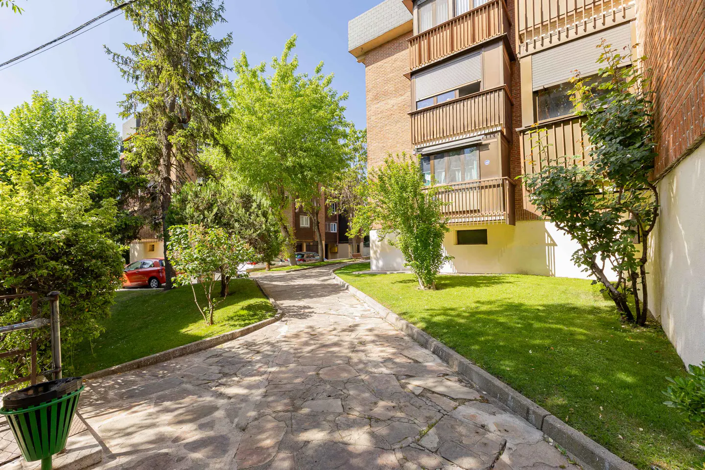 Exterior view of a brick apartment building with balconies, green lawn, trees, and a stone pathway.