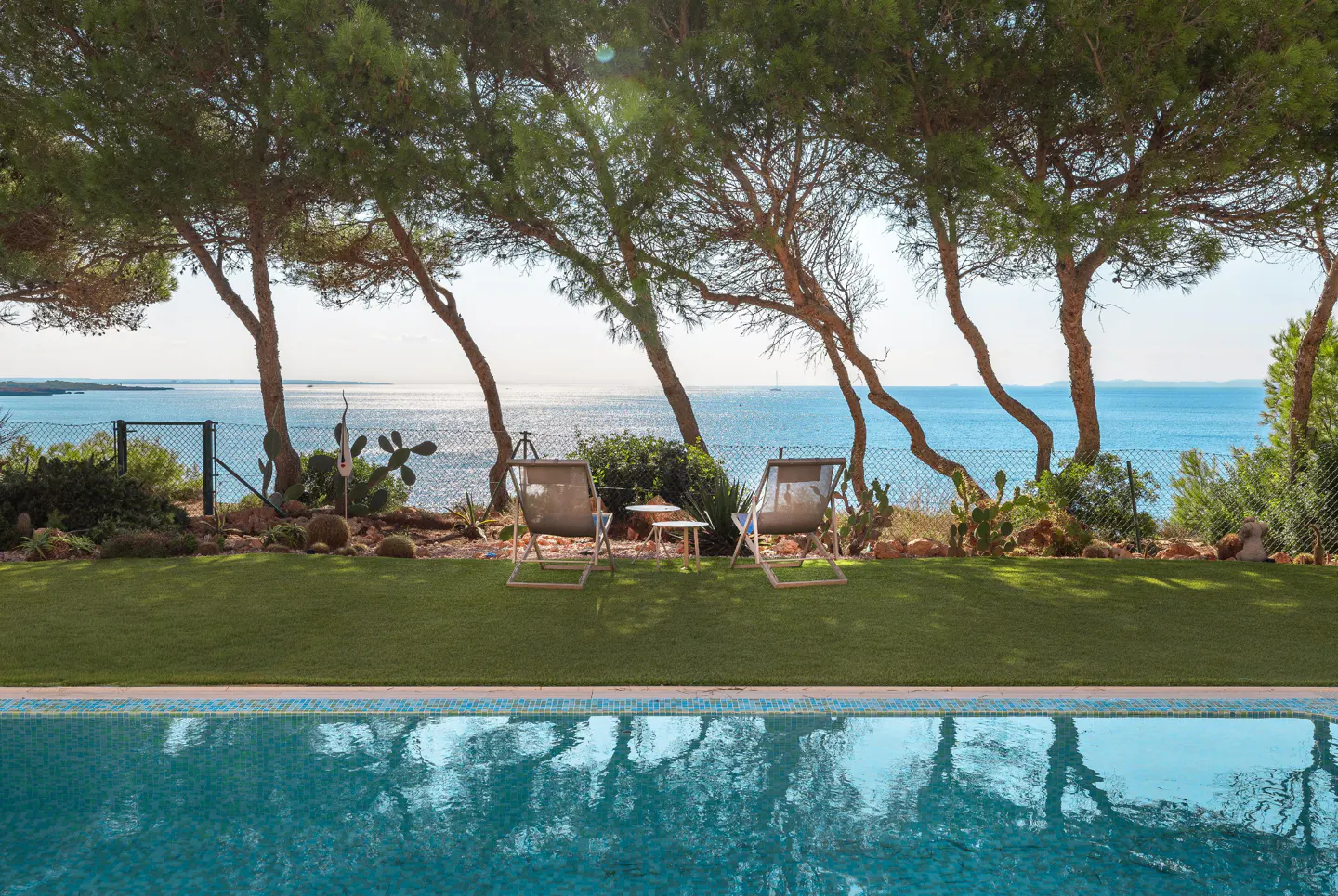 Poolside view with two chairs on green grass overlooking the ocean, framed by trees and a blue sky.