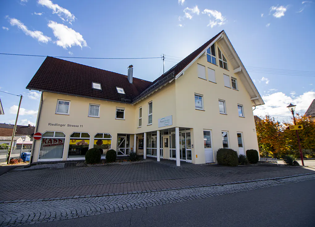 A two-story cream-colored building with a dark tiled roof, featuring ground-floor storefronts and a paved parking area.