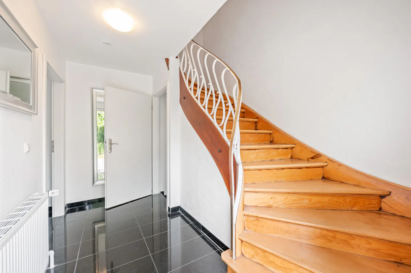 Bright entryway with black tile floor, white walls, and a curved wooden staircase with white railing. A white door is slightly ajar.