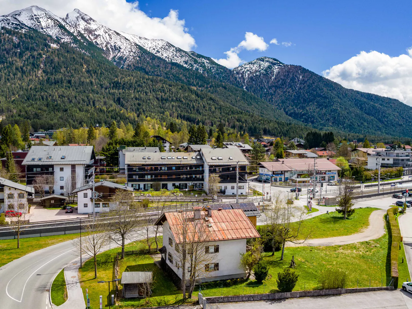 Scenic view of a European town nestled in the mountains, with snow-capped peaks, green trees, and clear blue sky.