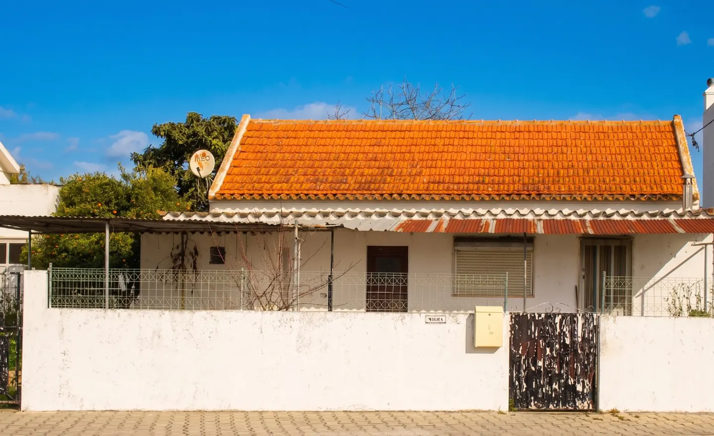 A white, single-story house with an orange tile roof, a white wall, and a black gate under a blue sky.