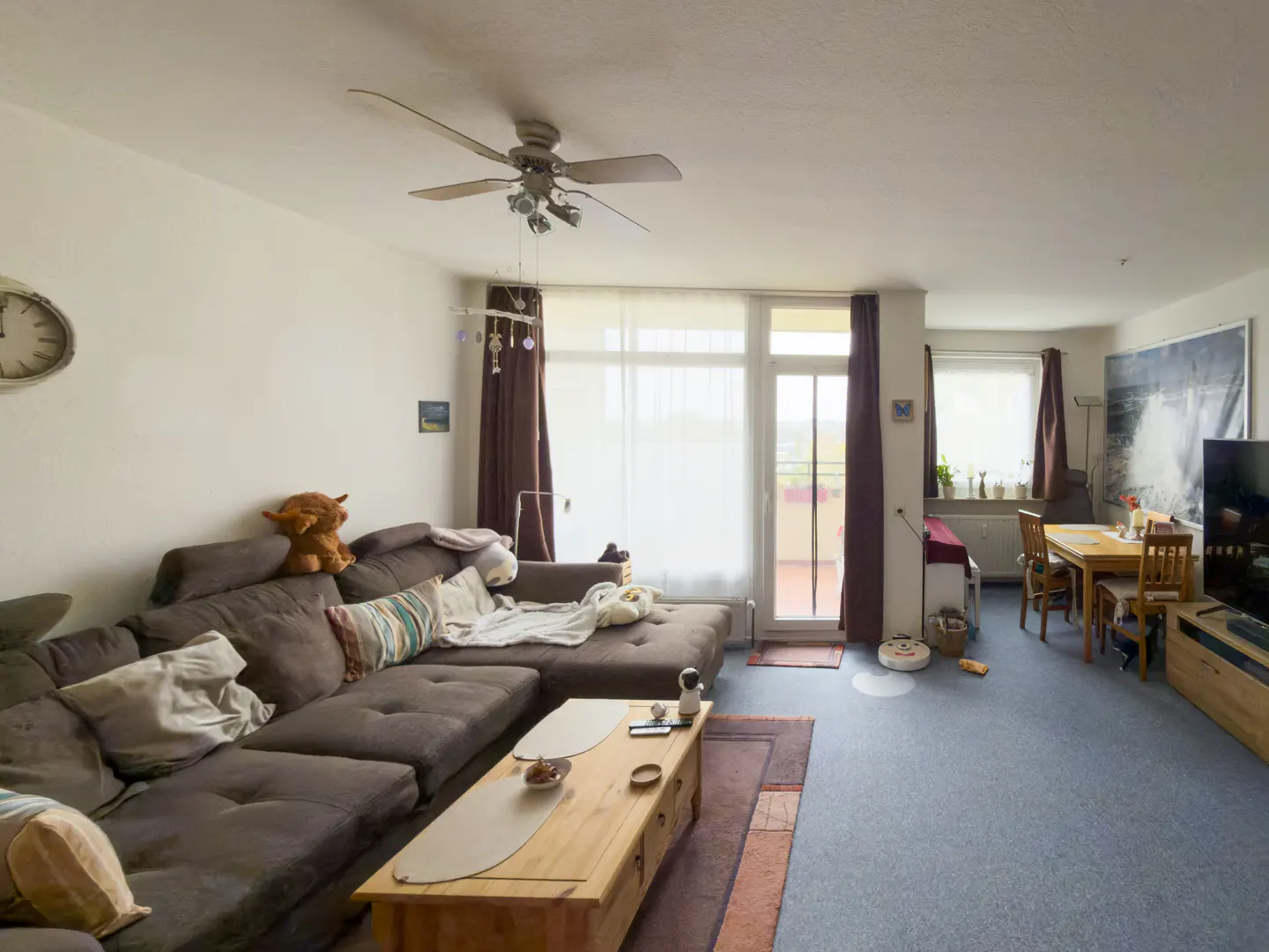 Living room with a large brown sectional sofa, wood coffee table, dining table, and balcony access. A ceiling fan hangs above.