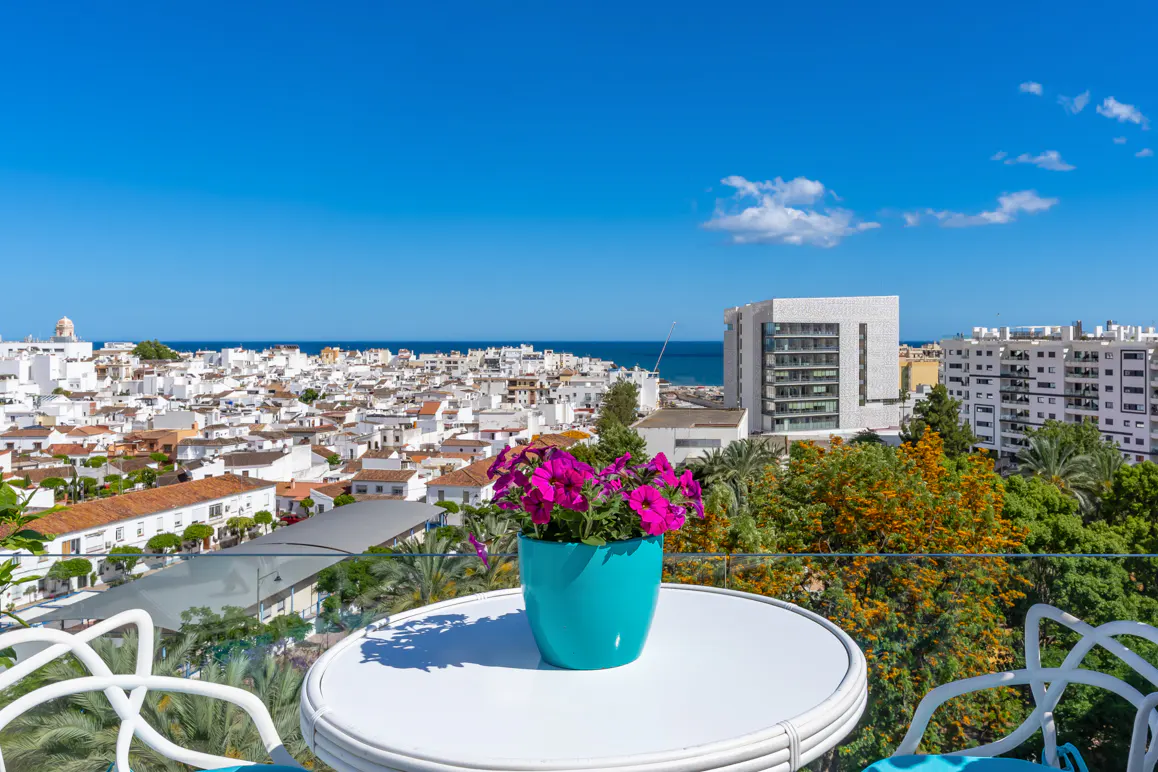 Balcony view with white table, chairs, and turquoise flower pot with pink flowers. Cityscape and ocean in the background under a blue sky.