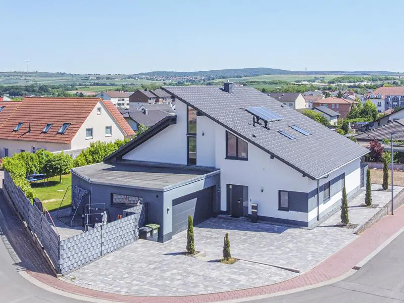 Aerial view of a modern white and grey house with a slanted roof, solar panels, and a paved driveway in a suburban area.