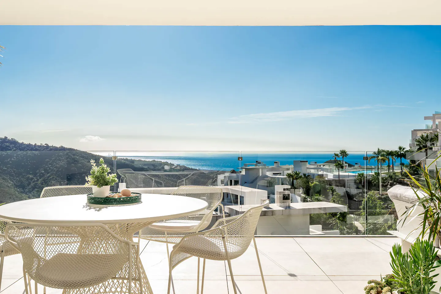 Balcony view with white table, chairs, and ocean backdrop. Blue sky, mountains, and modern buildings visible.