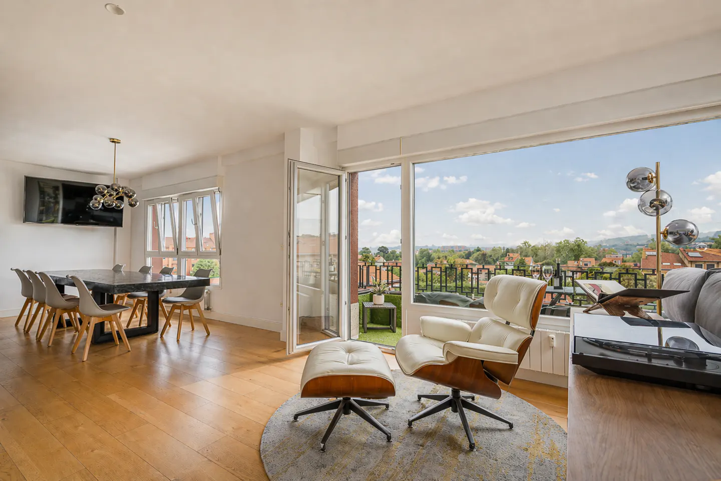 Bright, modern living room with wood floors, dining table, and a white chair with ottoman near a balcony overlooking a cityscape.
