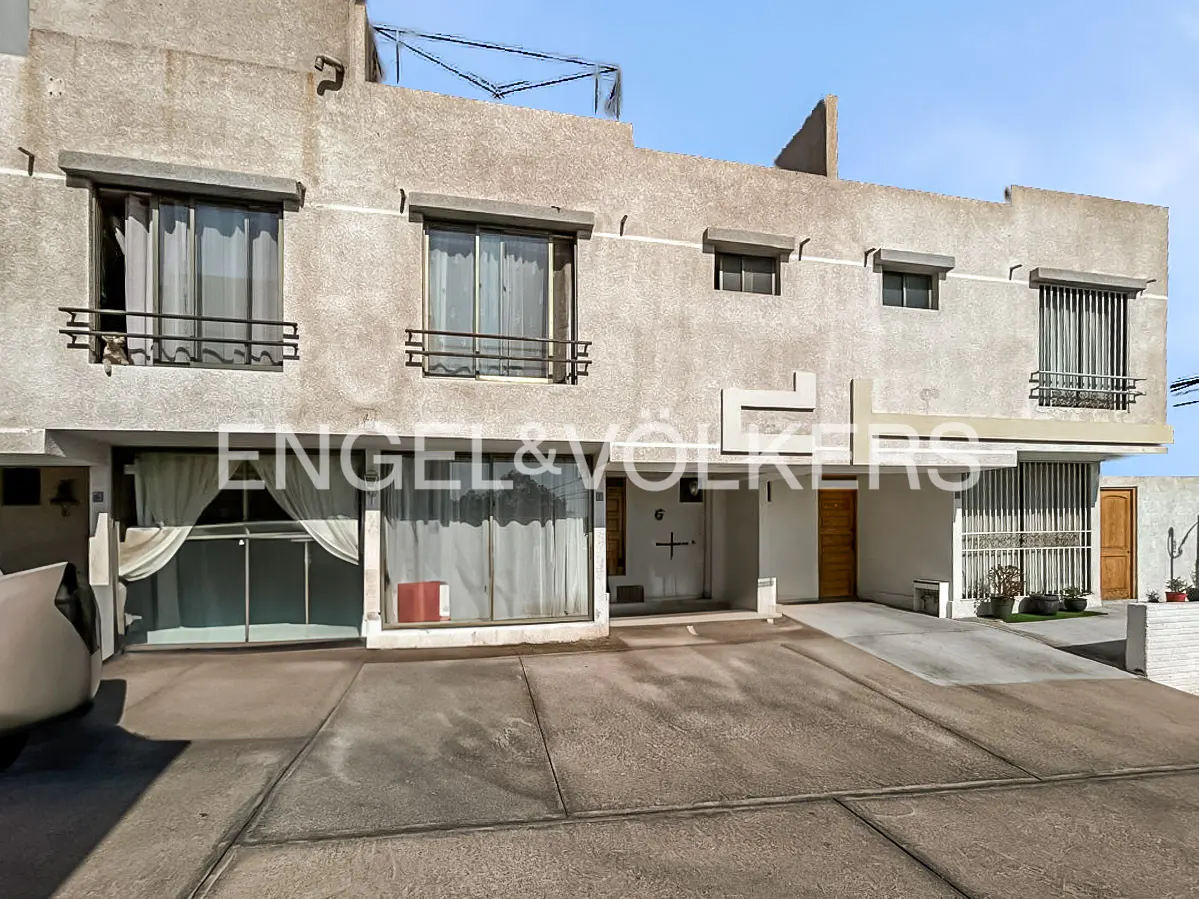 Exterior view of a three-story beige townhouse with large windows, curtains, and the Engel & Völkers logo. A concrete driveway leads to the building.