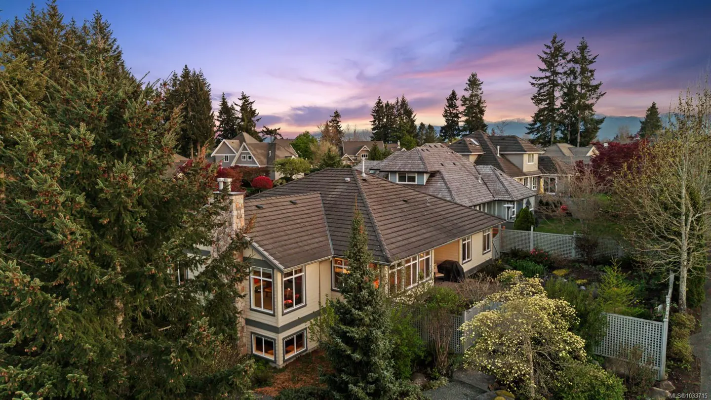 Aerial view of a beige two-story house with a brown roof, surrounded by green trees and other houses under a colorful sunset sky.