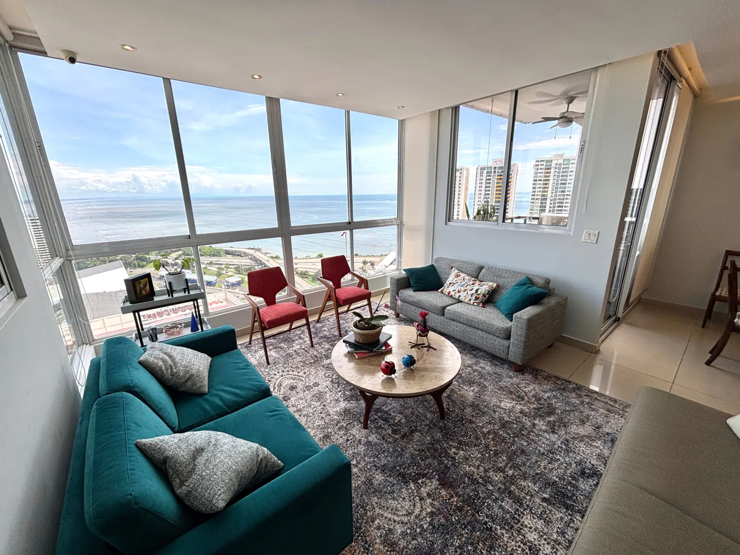 Bright living room with ocean view. Teal sofa, gray sofa, and red chairs surround a round coffee table on a patterned rug. Large windows overlook the ocean.