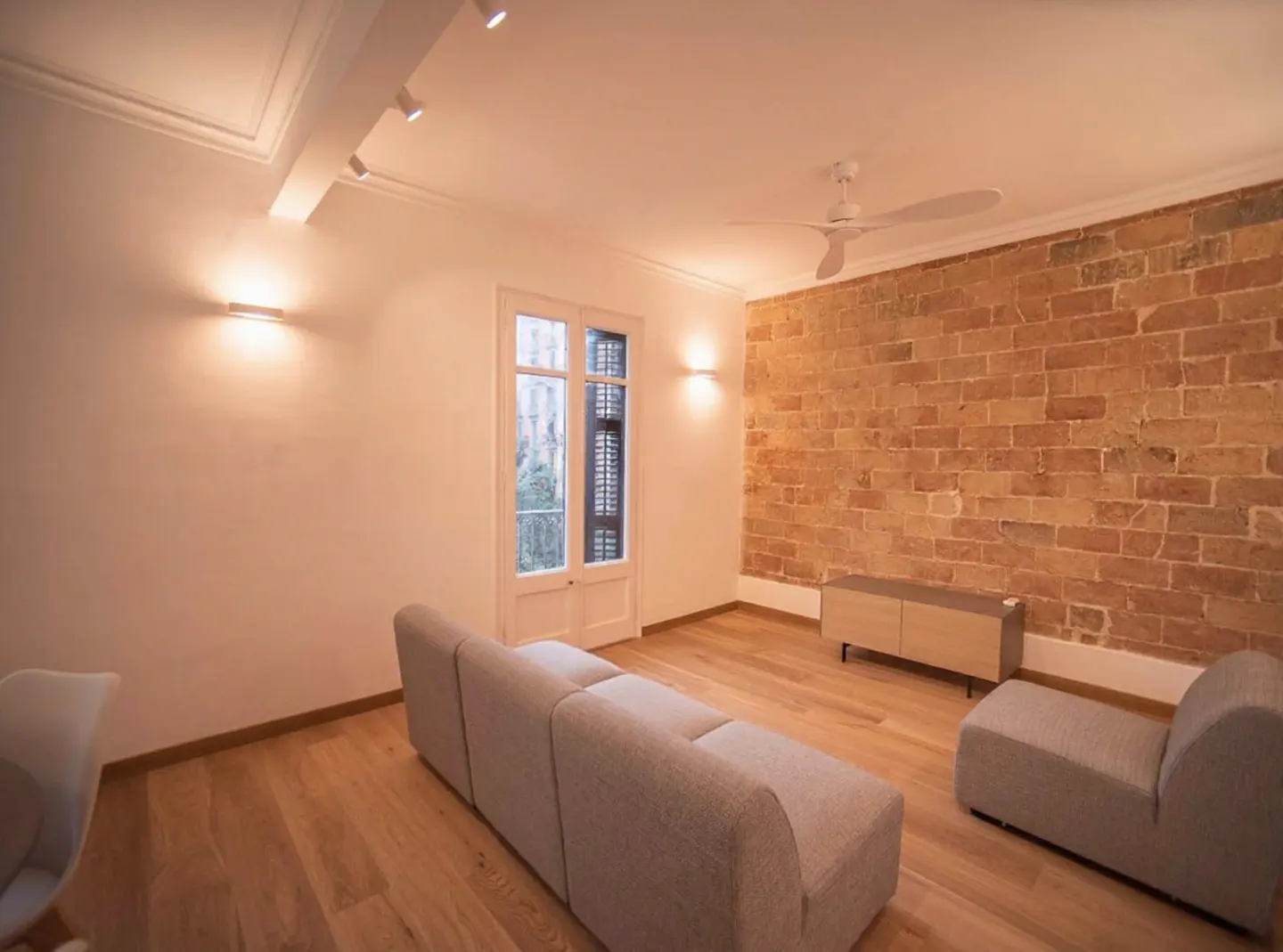 Bright living room with hardwood floors, a gray sofa, and a brick accent wall. A white ceiling fan and modern lighting fixtures are also visible.