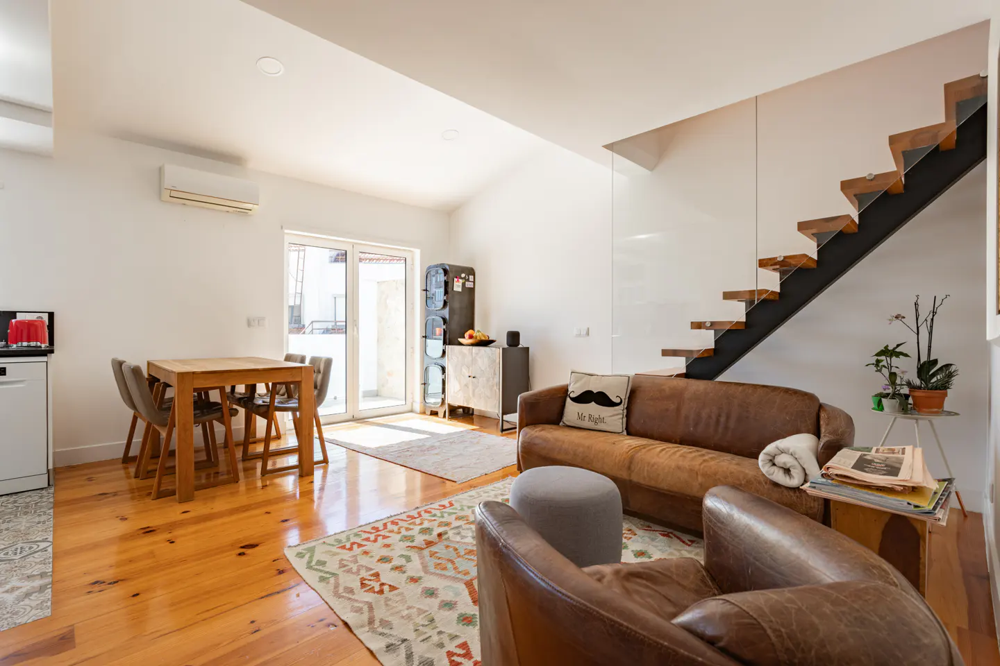 Modern open-plan living room with wood floors, brown leather sofa, armchair, dining set, and floating staircase.