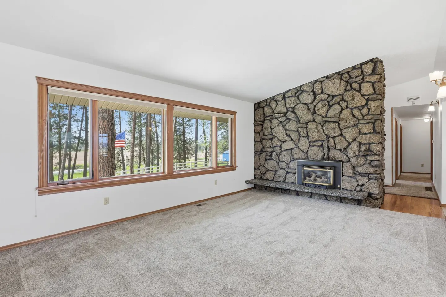 Living room with gray carpet, stone fireplace, and a window view of trees and an American flag.