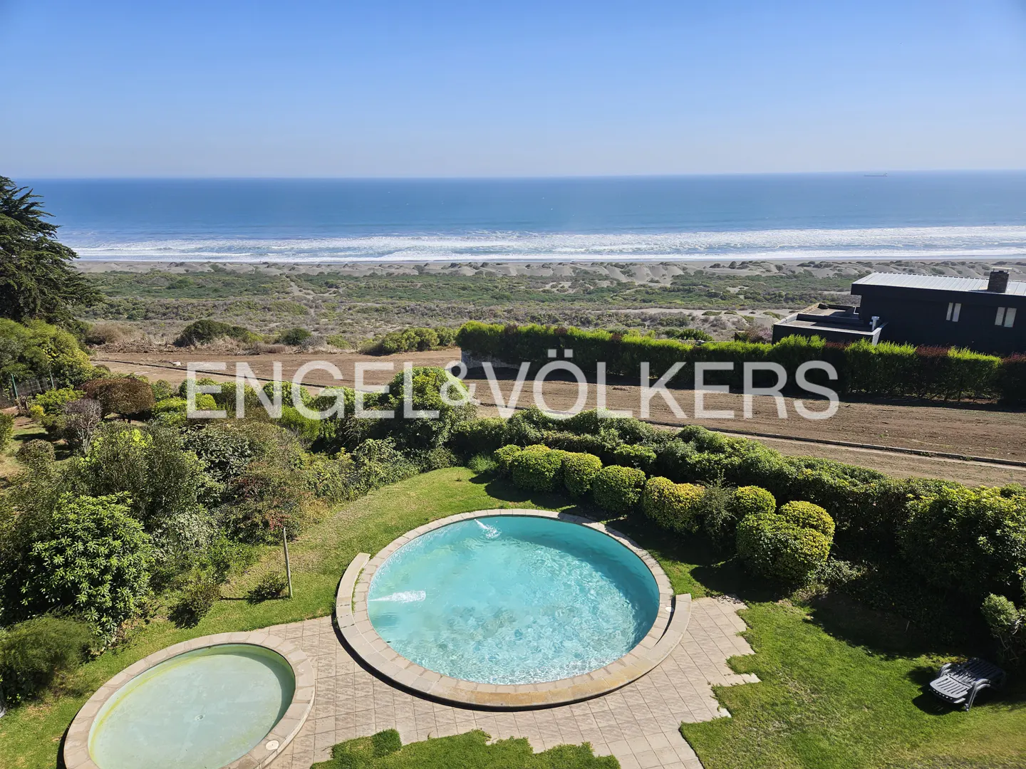 Aerial view of a round, turquoise pool in a green yard, with the ocean and beach in the background.