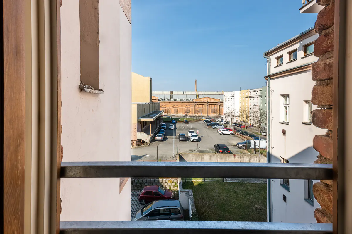 View from a window with a metal railing, overlooking a parking lot with cars and buildings in the background.