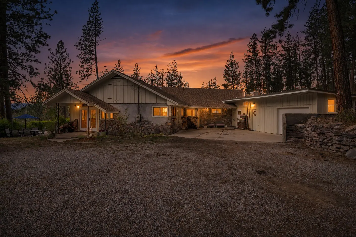Exterior view of a one-story house with a gravel driveway at dusk. The sky is a mix of orange and purple. Tall trees surround the house.