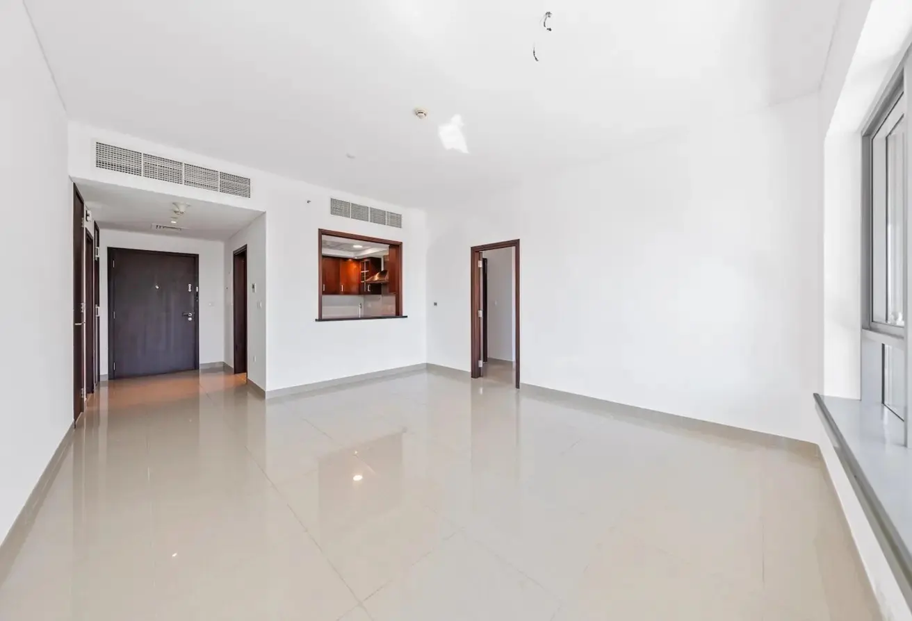 Bright, empty living room with white walls, beige tile floors, dark wood doors, and a kitchen pass-through window.