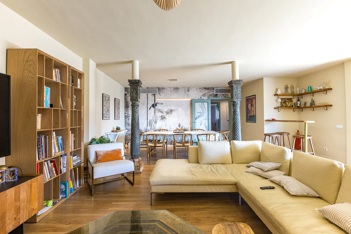 Bright living room with a cream sectional sofa, wood floors, and a dining area with a tropical mural. Bookshelves and bar area visible.