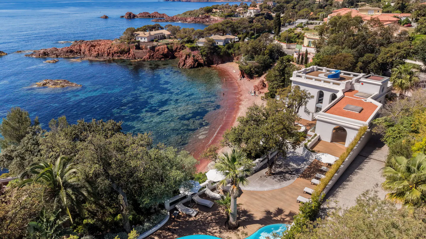 Aerial view of a white villa with a pool overlooking a red sand beach and turquoise water.