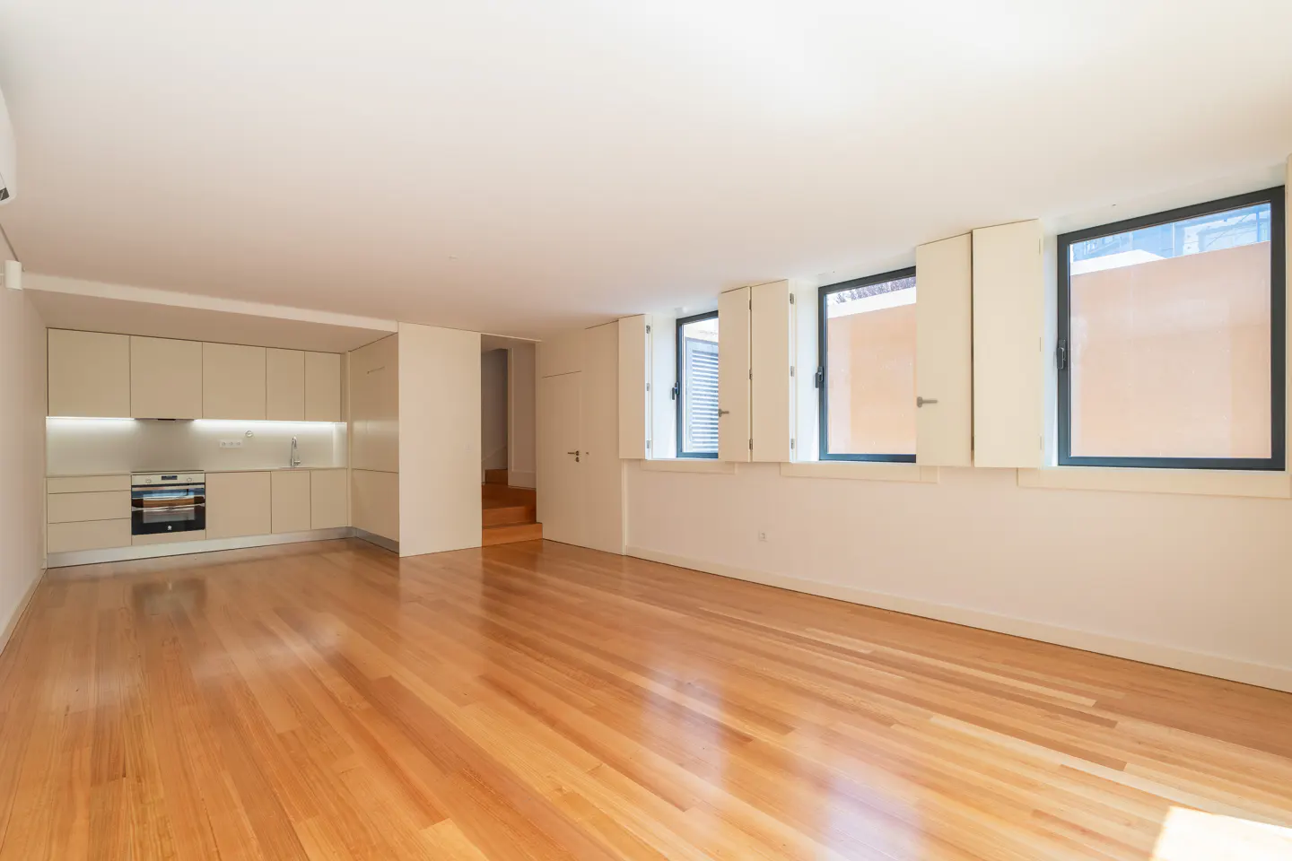 Bright, empty room with wood floors, white walls, and a kitchen area with light cabinets. Three windows with white shutters line the wall.