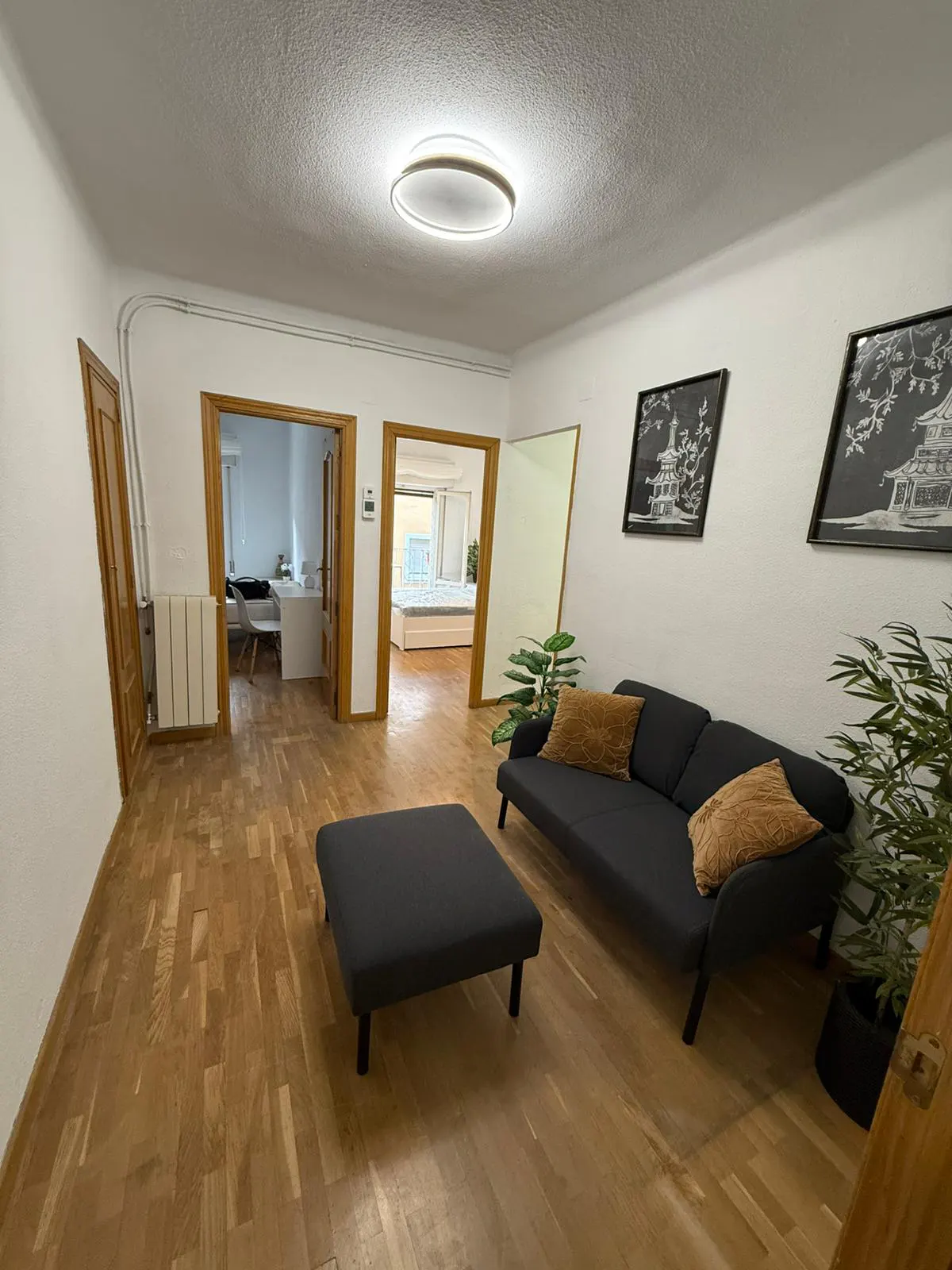 Living room with wood floors, white walls, and a black sofa with brown pillows. Two framed pictures hang above the sofa.