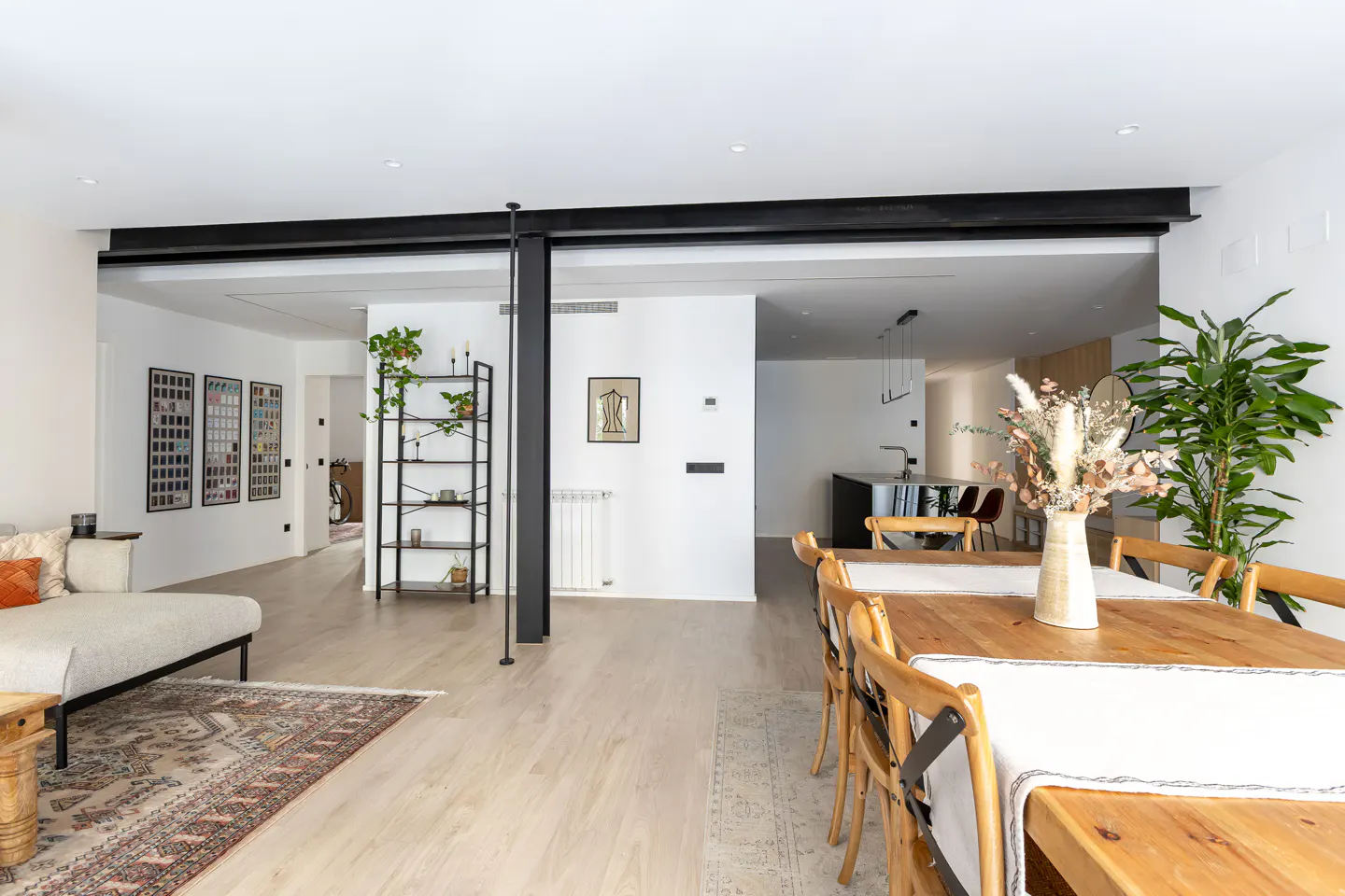 Open-concept living space with light wood floors, white walls, and black steel beams. A dining table and sofa are visible.