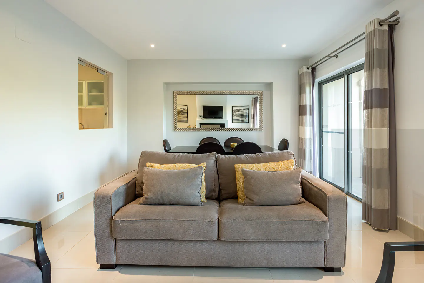 A grey sofa with yellow accent pillows sits in a bright living room, with a dining area and large mirror in the background.