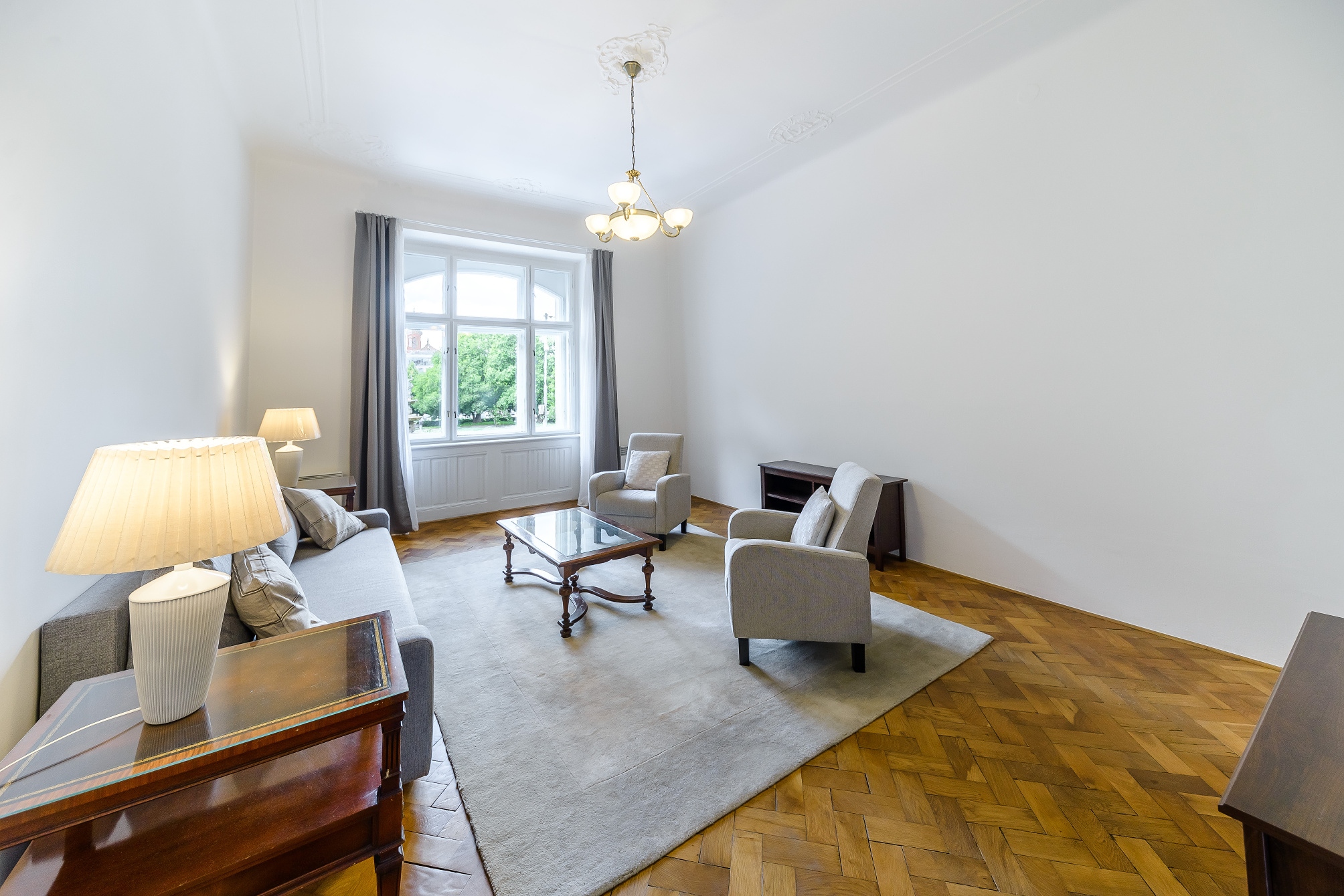 Bright living room with herringbone wood floors, a gray sofa, two armchairs, a glass coffee table, and a chandelier. [AI-generated text]