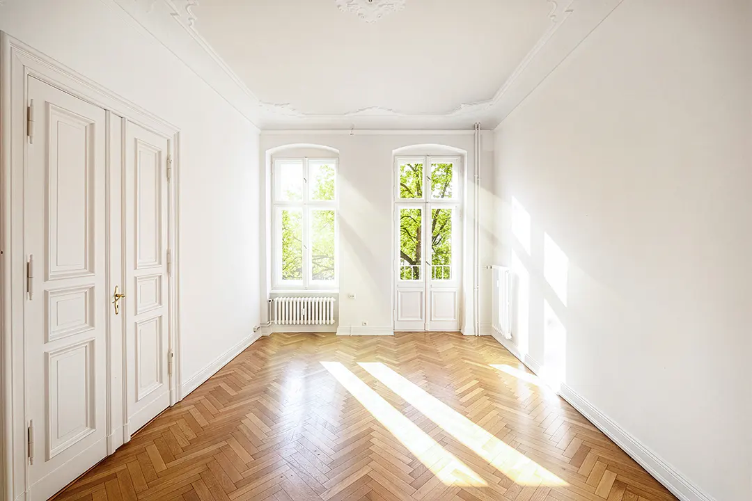 Bright, empty room with herringbone wood floors, white walls, and two windows showing green trees outside. A white door is on the left.